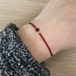 Può includere: Un braccialetto di filo rosso con una piccola gemma rotonda, rosso scuro e dettagli dorati. Il braccialetto è indossato su un polso, con una manica di maglione grigia e bianca sullo sfondo.