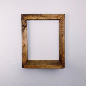 May include: A rustic, square wooden shelf with a warm brown stain. The shelf is mounted on a white wall and features a simple, open design. The wood grain is visible, adding to its natural aesthetic. Perfect for displaying small items.