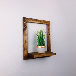 May include: A square, wooden floating shelf with a dark brown finish. The shelf holds a small, white pot with a gold rim, filled with green faux grass. The shelf is mounted on a white wall.