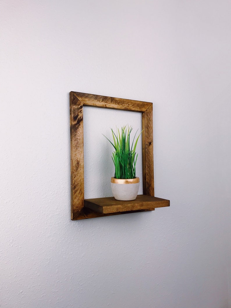 May include: A square, wooden floating shelf with a dark brown finish. The shelf holds a small, white pot with a gold rim, filled with green faux grass. The shelf is mounted on a white wall.
