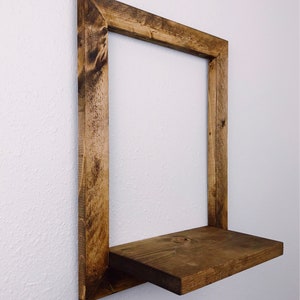 May include: A rustic, square wooden shelf with a dark stain. The shelf is mounted on a white wall and features a rectangular frame with a single shelf. The wood grain is visible, adding to the natural aesthetic.