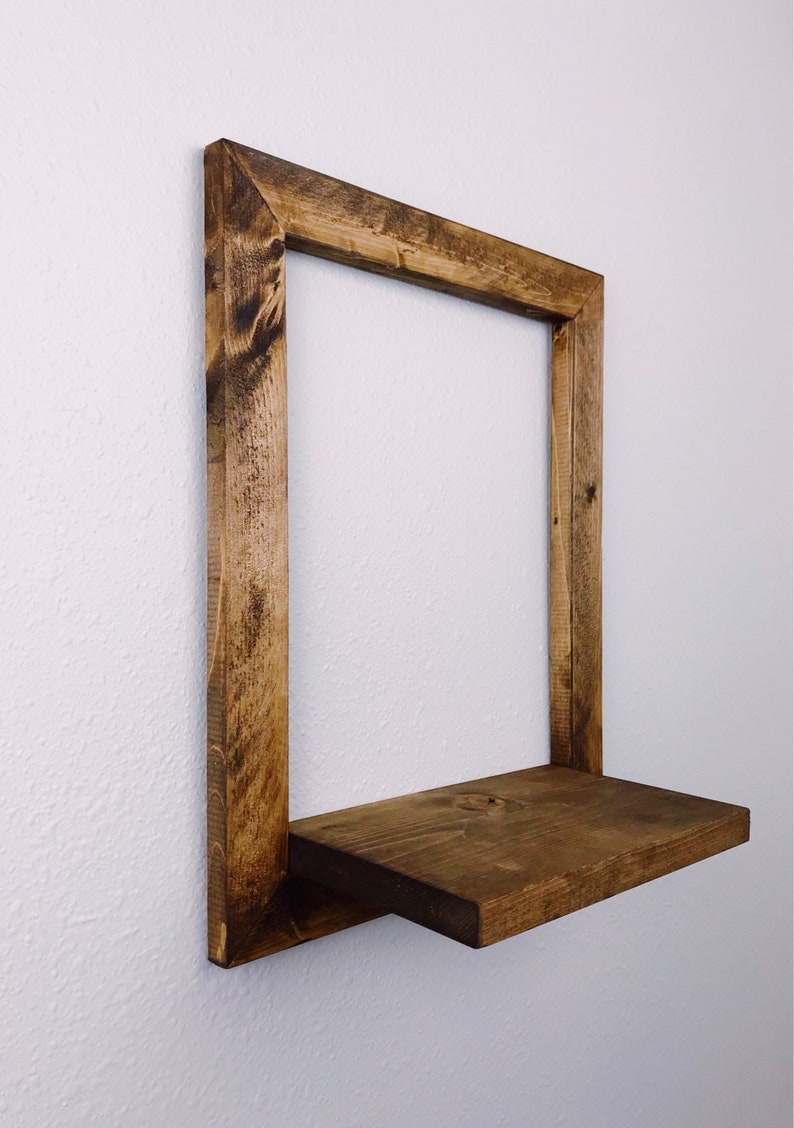 May include: A rustic, square wooden shelf with a dark stain. The shelf is mounted on a white wall and features a rectangular frame with a single shelf. The wood grain is visible, adding to the natural aesthetic.