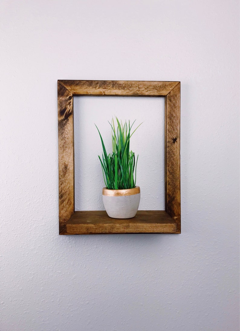 May include: A rustic, square wooden shelf with a live plant in a gray pot with a gold rim. The shelf is made of dark brown wood and is mounted on a white wall. The plant has long, green blades.