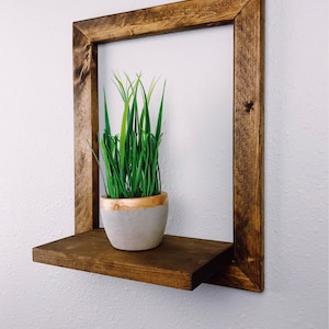 May include: A square, dark-stained wooden shelf with a lower shelf and a frame. A small, gray pot with a gold rim holds green grass. The shelf is mounted on a white wall, creating a minimalist, modern aesthetic.