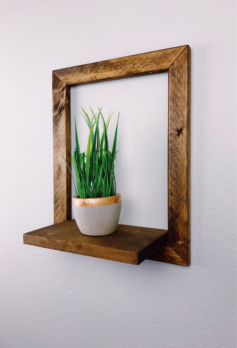 May include: A square, dark-stained wooden shelf with a lower shelf and a frame. A small, gray pot with a gold rim holds green grass. The shelf is mounted on a white wall, creating a minimalist, modern aesthetic.