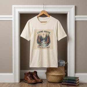 May include: A cream-coloured t-shirt with a nature scene depicting a river, trees, and a cross. The text on the shirt reads "Living Water" and "Never Thirst Again." The t-shirt is hanging in a doorway with brown boots, a basket, and books in the foreground.