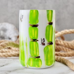 Puede incluir: Un vaso de cerámica blanco con un diseño de bambú verde y detalles de mariposas.
