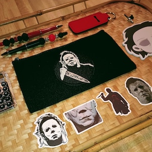 May include: A black zippered pouch with a white embroidered image of a character holding a knife and the text "No Lives Matter". The pouch is surrounded by various stickers featuring the same character in different poses. The stickers are on a woven bamboo surface.