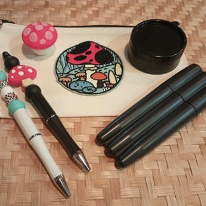 Può includere: Un assortimento di articoli a tema funghi, tra cui una gomma a forma di fungo rosso e bianco a pois, una toppa con un disegno di funghi, un contenitore nero e diverse penne nere. Sono inclusi anche una penna con perline e una penna bianca.