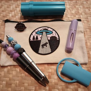 Könnte beinhalten: Eine weiße Canvas-Reißverschlusstasche mit einem schwarzen Reißverschluss und einem schwarzen gestickten Aufnäher eines UFOs mit dem Text "Don't Panic". Die Tasche liegt auf einer geflochtenen braunen Oberfläche mit einer silbernen Nadel, zwei Stiften, einer blauen Röhre, einem blauen Ring mit einem blauen Rechteck und einer violetten Flasche.