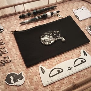 May include: A black canvas pouch with a white cat design, a black and white cat pin, a black and white cat ruler, a black and white cat sticker, and a black and white cat bookmark. The pouch is on a woven bamboo tray.