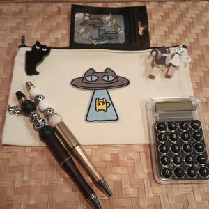 May include: A white zippered pouch with a black cat, a UFO with a yellow cat inside, and a calculator. The pouch is decorated with black and white beads and a gold pen.
