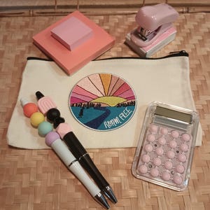May include: An assortment of office supplies including a pink stapler, calculator, sticky notes, and pens. A canvas pouch features a colorful embroidered patch with the text "ROAM FREE". The pens are black and white with silver accents. A beaded pen accessory is also included.