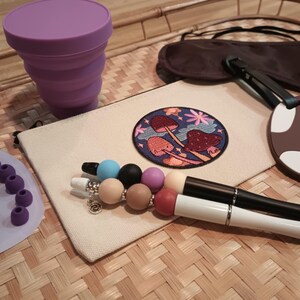 May include: A close-up of a set of travel essentials, including a collapsible silicone cup, a sleep mask, a mushroom-themed embroidered patch, a beaded bracelet, and two pens. The items are arranged on a woven surface.
