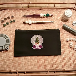 Puede incluir: Una bolsa negra con cremallera con un diseño de bola de nieve bordada en blanco. La bolsa está sobre una bandeja de bambú tejido con varios artículos pequeños, incluyendo un bolígrafo, una pequeña botella y algunos artículos decorativos.