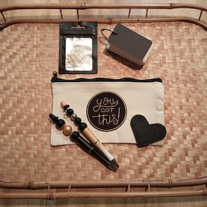 May include: A white canvas pouch with a zipper closure. The pouch has a black circle with gold lettering that says "You Got This!" and a black heart-shaped patch. The pouch is sitting on a woven bamboo tray with two black and gold pens, a small bag of gold paper clips, and a black notepad.
