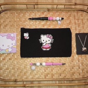 Könnte beinhalten: Eine schwarze Reißverschlusstasche mit einem Hello-Kitty-Stickmotiv. Die Tasche befindet sich auf einem geflochtenen Bambus-Tablett mit einem rosa und weißen Hello-Kitty-Notizblock, einem rosa Stift mit weißen Perlen, einem schwarzen Stift mit rosa und weißen Perlen und einer schwarzen Karte mit einer silbernen Hello-Kitty-Anhängerkette.