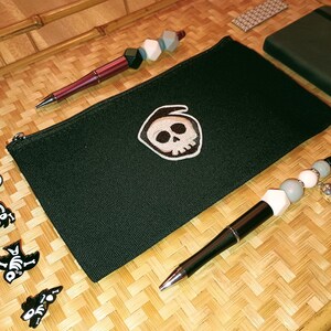 May include: A black zippered pouch with a white skull and crossbones design. The pouch is lying on a woven surface with a pen and a beaded pen with a skull charm.