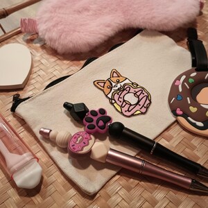 May include: A white canvas pouch with a pink and white embroidered corgi design, a brown and white donut-shaped keychain, a pink and white eye mask, a pink and white toothbrush holder, and two pens with pink and brown accents.