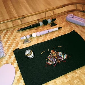 May include: A black zippered pouch with a colourful moth embroidered on the front. The pouch is on a woven bamboo tray with a calculator, two pens, and two heart-shaped notepads.