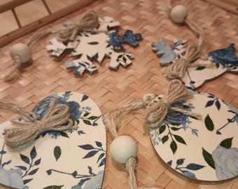 Op de afbeelding: Een set van vijf houten ornamenten met blauwe en witte bloemenpatronen. De ornamenten zijn gevormd als een hart, een sneeuwvlok, een cirkel en twee rendieren. Elk ornament heeft een touw strik en een touw om op te hangen.