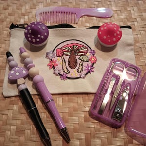 Puede incluir: Una bolsa de tocador de color morado y blanco con un diseño de bordado de hongos. La bolsa contiene dos pequeños recipientes con forma de hongo de color morado y blanco, dos bolígrafos morados con tapas de hongo, un peine morado y un juego de cortaúñas morado.