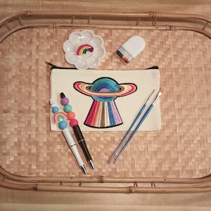 May include: A white canvas pouch with a rainbow embroidered planet design. The pouch is on a woven tray with two paintbrushes, a paint palette, and a pen with rainbow beads.