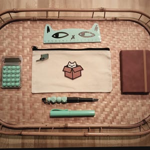 Puede incluir: Una regla en forma de gato azul claro, una bolsa de lápices blanca con un diseño de gato en una caja, una calculadora azul claro, una libreta marrón, un bolígrafo azul claro y un bolígrafo negro con un llavero de cuentas azules claros están dispuestos en una bandeja de bambú tejido.