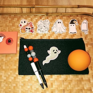 May include: A black zippered pouch with a ghost patch, two pens, a beaded keychain, and a bright orange ball. There are also several ghost-themed iron-on patches on a woven tray.