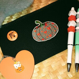 Puede incluir: Estuche de lápices de lona negro con un diseño de calabaza blanca y naranja, un pin de telaraña y dos bolígrafos con tapas de colores.