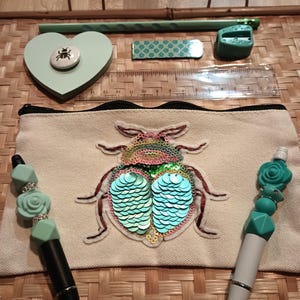 May include: A white canvas pencil case with a green and blue sequined beetle design. The case is open and has a black zipper. There are two pens inside the case, one with a green and blue rose design and the other with a black and white design.