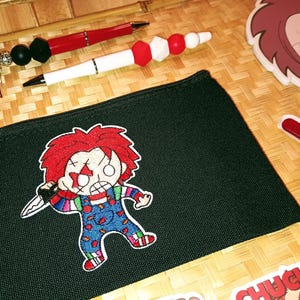 May include: Black zippered pencil case with a red and blue embroidered cartoon character holding a knife. The character has red hair and a white face with a red scar. The character is wearing a blue and red striped shirt and blue overalls.