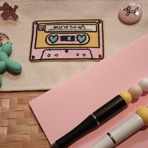 Może przedstawiać: Biała płócienna kosmetyczka z naszywką w postaci haftowanej kasety magnetofonowej w kolorze różowym i żółtym z napisem "Best of the 80's". Jest też zielony brelok w kształcie zwierzęcia z balonów, różowy guzik z białym obramowaniem przedstawiającym zwierzę z balonów oraz dwa długopisy z kolorowymi koralikami.