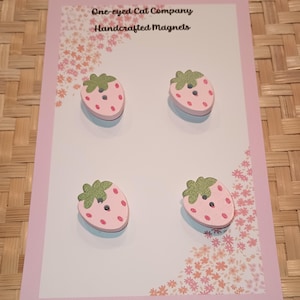 May include: Four pink and green wooden strawberry magnets. The magnets are on a white card with pink flowers and the text "One-eyed Cat Company Handcrafted Magnets".