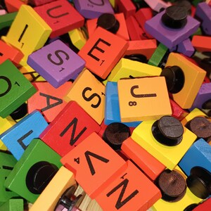 May include: A pile of colorful wooden letter tiles with black lettering and black plastic push pins. The tiles are in various colors, including red, green, blue, yellow, orange, and purple. The letters on the tiles include O, T, S, U, J, N, Z, A, E, and L.