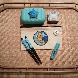 May include: A light blue tray with a light blue soap dish with a sparkly blue star, a small light blue bottle, a white zippered pouch with a yellow moon and blue clouds embroidered on it, and a small box with a floral design and the text "ENGLISH ROSE" on it.