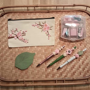May include: A bamboo tray with a white canvas pouch with pink floral embroidery, a clear plastic container with pink office supplies, and three pens with pink floral embellishments.