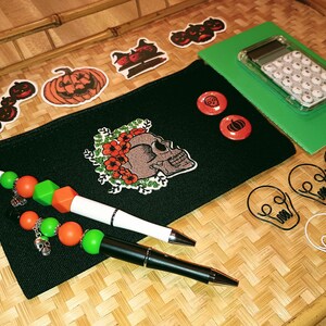 May include: A black zippered pouch with a skull and flowers embroidered on it. The pouch is surrounded by Halloween-themed stickers, including pumpkins, cats, and skulls. There are also two pens with colorful beads, a calculator, and skull-shaped paper clips.
