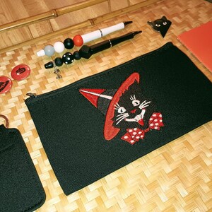 Può includere: Borsa nera con cerniera con un gatto nero che indossa un cappello da strega e un papillon rosso e bianco a pois ricamato sul davanti. La borsa è su una superficie di bambù intrecciato con altri oggetti a tema Halloween.