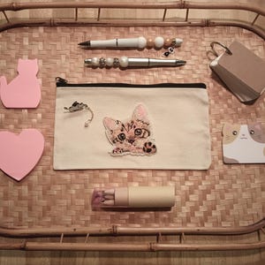 Könnte beinhalten: Ein weißer Stoff-Bleistiftetui mit einem gestickten braunen und weißen Kätzchen auf der Vorderseite. Das Etui liegt auf einem geflochtenen Bambus-Tablett mit rosa und braunen Haftnotizen, einem Stift und einem braunen Notizblock.