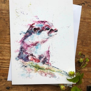 Puede incluir: Pintura acuarela de una nutria con tonos rosados, azules y verdes. La nutria está sentada en una roca y mirando hacia la derecha. El fondo es un lavado de color blanco.