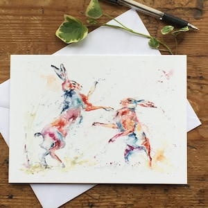 May include: Watercolor painting of two hares in a playful pose, with splashes of pink, blue, and yellow paint.