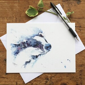 May include: A watercolor painting of a badger with blue and white tones. The badger is looking to the right and has a white stripe down its face. The background is white with a few blue watercolor splatters.