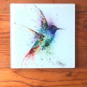 May include: Square glass coaster with a watercolour hummingbird design. The bird is rendered in shades of blue, green, purple, and red, with a splattered paint effect. A signature is visible in the bottom right corner.