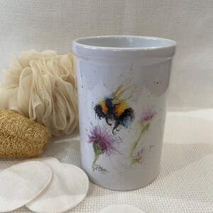 Könnte beinhalten: Weißer Keramikbecher mit einem Aquarellgemälde einer Hummel und lila Distelblüten. Der zylindrische Becher hat eine glatte, glänzende Oberfläche. Das Design zeigt schwarze, gelbe und orangefarbene Details. Wattepads und ein Luffa befinden sich im Hintergrund.