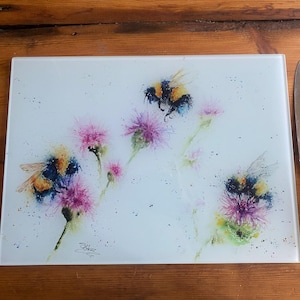Puede incluir: Tabla de cortar rectangular de vidrio con una pintura de acuarela de abejorros y flores de cardo morado. Un cuchillo de acero inoxidable con mango negro descansa a la derecha sobre madera.