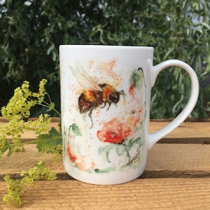 May include: White ceramic mug featuring a watercolour design of a bumblebee flying near a red poppy flower. The mug has a handle.