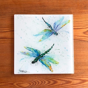 May include: Square glass coaster with a watercolour painting of two dragonflies. The dragonflies have blue and green wings with yellow accents, set against a white background with blue and green paint splatters. The coaster is on a wooden surface.