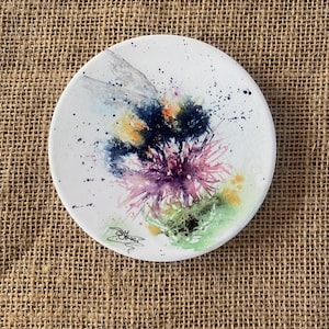 May include: A watercolor painting of a bumblebee on a pink thistle flower. The painting is on a white ceramic coaster with a green background and black splatters.