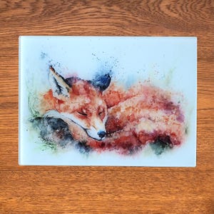 Puede incluir: Una pintura de acuarela de un zorro rojo durmiendo en un lecho de hierba verde. El zorro está enroscado con la cabeza apoyada en sus patas. El fondo es de un azul suave con salpicaduras de pintura blanca.